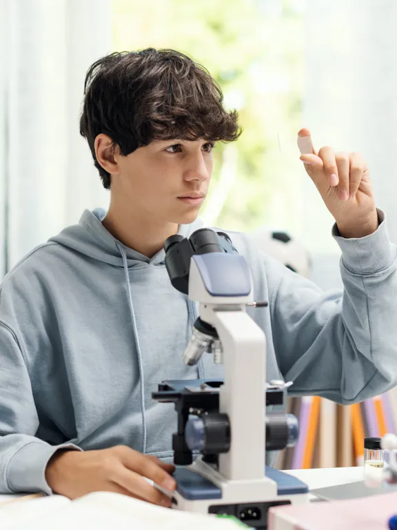 Student examining a specimen through a microscope at a desk in a bright science classroom